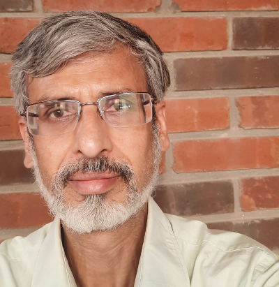 Man with glasses, gray hair, and a beard wearing a light shirt, standing against a brick wall.